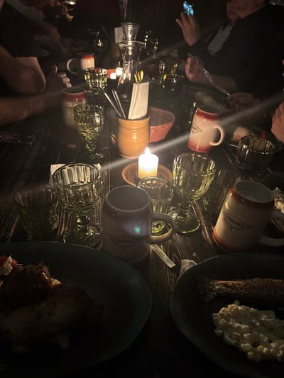 The image captures a dimly lit interior scene within the U Pinkasů establishment in Capital City of Prague, Czechia. At least five individuals are seated around a dark, distressed wooden table, their upper bodies and arms partially visible. The scene is illuminated primarily by a central white candle in a ceramic dish, casting a warm glow and producing lens flares. The table is set with multiple patterned, green-tinted glass goblets and tumblers, alongside traditional ceramic beer mugs, some bearing the "U Pinkasů" logo. A tall, light brown ceramic pitcher holds white napkins and various cutlery. A clear glass carafe stands near the center. Food is present on dark teal plates; one plate in the foreground left contains pieces of meat and a red side, while a plate in the foreground right holds a whole cooked fish and a white, lumpy side dish. One person in the upper right is holding a mobile phone, its screen emitting a bright light. A small paper slip displaying "210 CZK" is visible on the table. The environment is rustic, consistent with a traditional Czech pub or restaurant.
