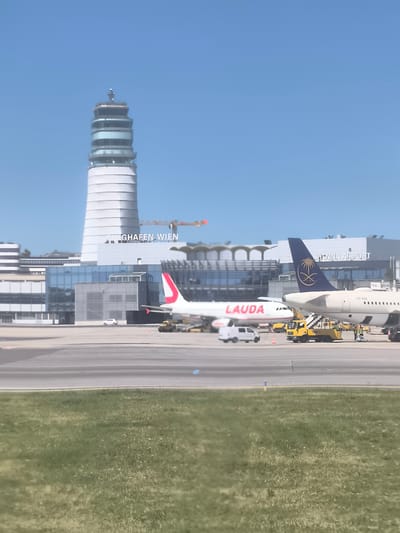 The image shows the Vienna International Airport in Schwechat, Austria, on a clear day. The airport's control tower and main terminal building are visible in the background. Two airplanes are parked on the tarmac: one with the "LAUDA" logo and the other with Saudi Arabian Airlines livery. Ground service vehicles and personnel are present around the airplanes. In the foreground, there is a patch of green grass.