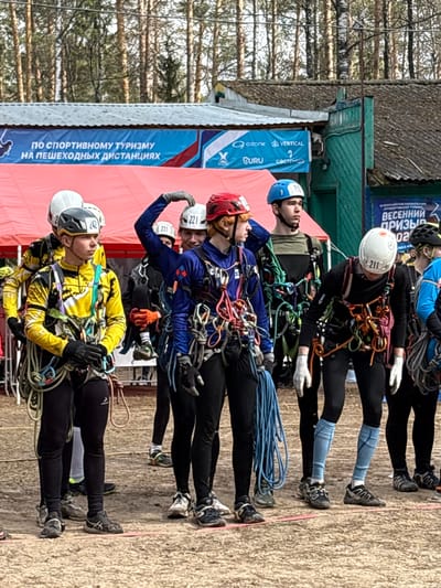 The image shows a group of seven people standing in a line outdoors, likely in Lesnoye, Russia, given the Cyrillic text on the banners visible in the background. They are all wearing helmets and climbing harnesses with ropes and carabiners, suggesting they are participating in a climbing or mountaineering event. The individuals are dressed in various athletic clothing, including yellow, blue, and black outfits. Each person has a number on their helmet. The ground appears to be dirt or packed earth. Behind the group are trees and structures, including a banner that reads "По спортивному туризму на пешеходных дистанциях" and "ВСЕРОССИЙСКИЕ СОM ПО СПОРТИВНОМУ ТУРЦИИ" which are Russian for "hiking distances" and "All-Russian competition in sports tourism," respectively. The overall scene conveys the setting of an outdoor sporting competition.