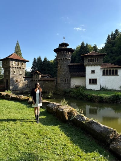 An outdoor, eye-level, full-shot photograph taken on a clear, sunny day, depicting a woman standing in a grassy area in front of the Château de Birtrange in Wal, near Groussbus, Luxembourg. The woman, an adult with dark hair, is positioned in the left mid-ground, wearing a light-colored dress, black blazer, black lace-up boots, and sunglasses; she is looking to her right. Behind her, a calm, murky green pond separates the foreground grass from the multi-structured stone and white-plastered château. The architectural complex features two prominent towers: a square stone tower with a red-tiled roof on the left, and a taller, cylindrical stone tower with an ornate circular roof and weather vane on the right. These are connected by lower stone walls and an adjacent white building with brown roofs, featuring arched and rectangular windows. Dense evergreen forest covers the hillside behind the château. Large, moss-covered stones line the edge of the pond in the mid-ground, with distinct shadows cast on the grass by the sunlight.