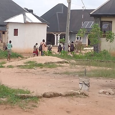 The image shows a dirt road in Adanisievbe, Nigeria (latitude: 5.955, longitude: 5.704). There is a group of children in school uniforms walking away from the camera down the road. In the foreground, there is a white bucket and some rocks. There are a few buildings in the background, some with black roofs and some with white roofs. There is also some foliage to the left and right of the road. The sky is overcast.