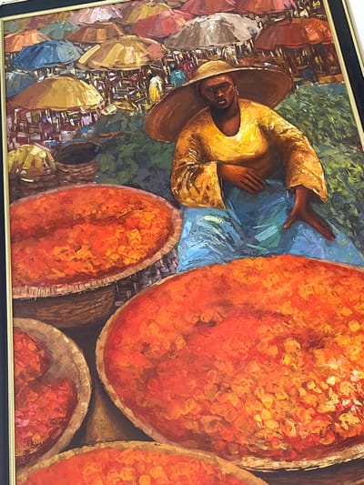 This oil painting depicts a vibrant outdoor market scene located in the city of Alaguntan, Nigeria. In the foreground, a dark-skinned individual, wearing a broad-brimmed straw hat, a bright yellow long-sleeved tunic, and a blue lower garment, is seated with a direct gaze forward. Their right hand rests on their lap, while the left hand is extended to the side. Positioned prominently around this figure are several large, round, woven baskets filled with a copious amount of vivid orange-red granular material, consistent with a foodstuff such as dried peppers or spices. The mid-ground and background reveal a bustling market ambiance characterized by numerous colorful umbrellas and canopies in shades of red, orange, brown, blue, and white, providing shade over indistinct market stalls and numerous background figures. The ground beneath the market appears to be a mix of earthy tones and green elements, indicative of an open-air setting. The entire composition is rendered with thick, visible impasto brushstrokes.