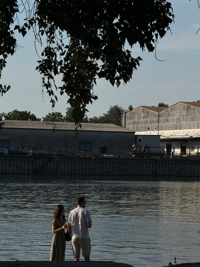 A man and a woman stand on a dock next to a body of water. The man is wearing a white t-shirt and tan shorts, and the woman is wearing a green dress. They appear to be conversing, and the woman is holding a glass. In the background, there are several large industrial buildings. The sun is low in the sky, casting long shadows, suggesting it is either early morning or late afternoon. The water is calm with slight ripples reflecting the sunlight. The overall scene suggests a peaceful, possibly romantic, moment by the water. The location is identified as Frankfurt, Germany. There is some graffiti visible on the side of one of the buildings.