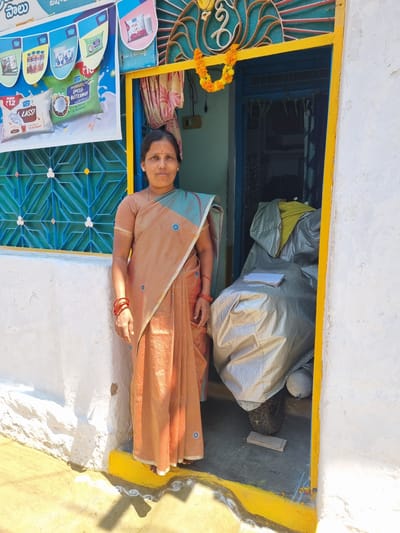 A full-length, front-facing image shows an adult Indian woman standing in a sunlit doorway in Pedda Kammavaripalli, India. The woman, appearing to be in her mid-forties, wears a light peach-colored striped saree with blue accents and small blue circular motifs, draped over a matching short-sleeved blouse. She has red bangles on both wrists, a small red bindi on her forehead, and a subtle smile. Her dark hair is pulled back.
To her left, a white textured wall features a light blue window covered by a geometric pattern grill. Above and partially obscuring the window, a multi-colored banner advertises "Hatsun Daily" dairy products, including Lassi and Buttermilk, with product images, Telugu text, and "MRP ₹12" visible.
The doorway is framed by a bright yellow outer border and a blue inner frame. Above the blue frame, an ornate teal and red metal grill is visible, beneath which a marigold garland hangs. Inside the doorway, a motorcycle or scooter is mostly covered by a silver tarpaulin, with a stack of white papers resting on top. A yellow fabric is visible near the top of the covered vehicle. A black tire and an exhaust pipe are partially visible beneath the cover, with a small wooden block on the dark concrete floor.
To the woman's right is a continuation of the white textured wall. The ground directly outside the doorway is light-colored concrete or packed earth, bordered by a yellow stripe at the base of the wall, with a faint white rangoli pattern visible. The scene is brightly lit by direct sunlight, casting distinct shadows.