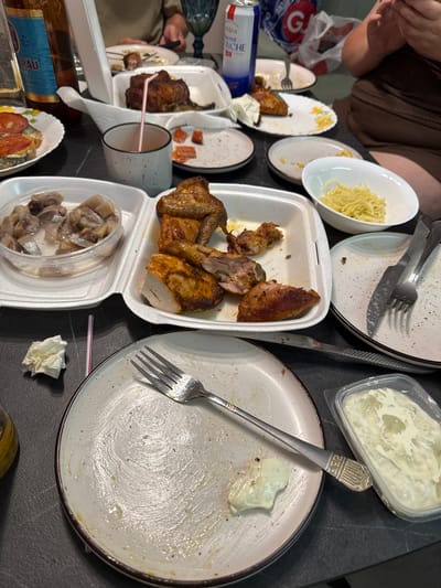 The image depicts a close-up view of a table with remnants of a meal, suggesting a post-dining scene. The table surface is dark, likely a countertop or table, and appears to be in an indoor setting. Various plates, some containing leftover food and others scraped clean, are scattered across the surface. There are also utensils, a glass of what appears to be beer, and a plastic container with a creamy substance.

Specific items visible include:
*   **Food remnants:** Pieces of cooked chicken, pasta-like strands, and a dish with sliced tomatoes and zucchini are present.
*   **Utensils:** Forks and knives are visible, some resting on plates, others on the table.
*   **Tableware:** A white styrofoam container holds chicken pieces. A white ceramic bowl contains the pasta. A small grey ceramic cup with a straw sits on the table. A glass bottle with a blue label, possibly beer, is partially visible on the left.
*   **Other items:** A crumpled white napkin is on the table. A small clear plastic container holds a light green creamy dip or sauce.

The overall impression is one of a casual, perhaps family or friend gathering, where a meal has recently concluded. The presence of a beer bottle and the style of the food suggest a relaxed atmosphere.

There is no information or visual evidence within the image to suggest this scene is located in the city of Novorossiysk, Russia. The description is limited to the observable details of the photograph.