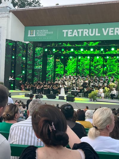 The image depicts an outdoor concert at the "Teatrul Verde" (Green Theatre) in Chișinău, Moldova. A large orchestra and choir are performing on a brightly lit stage, which is decorated with a backdrop of lush green foliage. In the foreground, an audience is seated on green benches, facing the stage. Several members of the audience are visible, including a woman with blonde hair tied in a ponytail, a person with gray hair and a striped shirt, and a person with dark hair and a black top. The stage banner above the performance displays the text "PRIMĂRIA MUNICIPIULUI CHIȘINĂU" (Chisinau Municipality Mayor's Office) and "TEATRUL VER" (Green Theatre). The overall atmosphere appears to be one of a cultural event or festival.