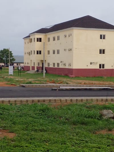 The image shows a modern, two-story building with a dark tiled roof, painted in cream and maroon. Several windows are visible on both floors, some open and some closed. There is a sign with "tetfund" printed on it on the right side of the building. The building is located in a grassy area with a paved road in the foreground. Several people are visible near the building, some walking and some sitting. In the background, there are other buildings and trees. The sky is overcast. The scene appears to be a public institution or a residential complex. The overall impression is one of a developing urban environment.