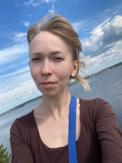 A selfie-style portrait captures a young adult female outdoors at what appears to be Пятый километр, Russia. The image is rotated 90 degrees clockwise, orienting the sky to the left and water to the right. The woman, with light skin and blonde hair tied back with some loose strands, looks directly at the camera with a neutral to slight smile. She has light-colored eyes and a few freckles or small marks visible on her face. She wears a dark brown long-sleeved top, and a prominent bright blue strap, possibly from a bag, crosses her chest. A gold-toned ear cuff is visible on her right ear.

The background is split between a clear blue sky with scattered white cumulus clouds on the left and a wide expanse of calm, dark blue water on the right. Along the distant horizon line across the water, faint traces of land with some structures or vegetation can be seen. A small portion of green foliage, likely a bush or tree, is visible in the bottom right corner of the frame. The bright lighting suggests it is daytime with sunny weather. There is no visible text in the image.