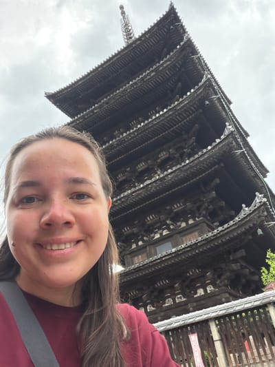 The image displays a female subject positioned in the left foreground, smiling directly at the camera. She has light skin, long brown hair, and is wearing a maroon long-sleeved shirt with a gray strap diagonally across her chest. The background is dominated by the Goju-no-to (Five-story Pagoda) of To-ji Temple, located in Minami Ward, Kyoto, Japan. The pagoda is constructed of dark wood, featuring multiple tiers with prominent eaves and a slender finial at its apex. The sky above is overcast. In the lower right, a section of a traditional wooden fence is visible, behind which a red and white banner with Japanese text and some green foliage can be seen.