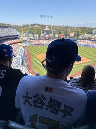 The image presents an elevated, over-the-shoulder view of a baseball game unfolding on a sunny day at an outdoor stadium. A clear blue sky stretches above, with distant green hills and light towers visible beyond the playing field.
The primary subject in the foreground is the back of a person's head and shoulders. This individual wears a blue baseball cap featuring a white heart and an MLB logo on its side, along with sunglasses perched on the back of their head. They are clad in a white jersey that prominently displays the name "大谷 翔平" (Ōtani Shōhei), referring to Japanese baseball star Shohei Ohtani, in large gold Japanese characters across the back. An MLB logo patch is also visible on the jersey's left shoulder.
To the left of this person, another spectator wears a blue baseball cap decorated with pink cherry blossoms. Further right, a person with blonde and brown hair, wearing a striped shirt, is partially visible. The stadium's seating sections are filled with numerous spectators, indicating a well-attended event.
The baseball field itself features a distinct dirt infield and green outfield, with players visible on the ground. In the background, beyond the field, a large video scoreboard and several light towers rise. Various blurry advertisements and signs are discernible along the stadium's perimeter, including text like "METRO" and "Scully & Scully." A small American flag can also be seen in the distance. The bright, clear conditions indicate a daytime game, and the presence of Shohei Ohtani's name, combined with the blue caps and the Los Angeles context, strongly suggests Dodger Stadium in Los Angeles, United States.