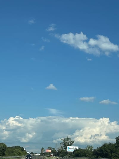 This media file captures a bright, clear sky with scattered white clouds, suggesting a pleasant daytime weather. The perspective is from a vehicle moving along a road, indicated by the slight blur and the roadside elements visible on the right side. These elements include a line of trees, a highway barrier, and a few billboards. One billboard is clearly visible and displays text that reads "mobile.bg" along with a blue icon. Another billboard further down the road shows a red and white advertisement. Cars are also visible on the road, indicating ongoing traffic. The overall impression is that of a typical travel scene on a sunny day. The scene appears to be located in Lozen, Bulgaria, given the provided location context.