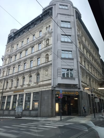 The image captures a street view in Prague, Czechia, featuring the corner of a multi-story building with ornate architectural details. The building displays cream-colored facades with numerous windows, some featuring decorative arched tops. The ground level of the building houses a Commerzbank branch, identifiable by its prominent signage and large glass windows. A pedestrian crossing is visible in the foreground, suggesting a busy urban intersection. Several utility poles and overhead wires are present, indicative of an established city infrastructure. The sky appears overcast, contributing to a muted, cool-toned lighting in the scene. A person is standing near the entrance of the Commerzbank.