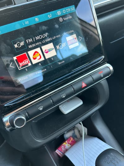 The scene depicts the interior of a vehicle, specifically the dashboard and center console area, located in Veauchette, France. A central infotainment screen, exhibiting minor dust and smudges, displays an active French radio tuner interface. The currently selected station is "FM / MOUV'" at 88.00 MHz, identified as "LE WAKE-UP MIX - L'INTEGRAL." Other visible radio station logos include "france inter," "radio SCOOP," "funradio," a second "MOUV'," and "SKYROCK." The screen also indicates a time of 09:32, along with various system icons.
Directly below the screen, a horizontal row of physical controls features a chrome-trimmed rotary knob on the far left. To its right are buttons for front defrost, rear defrost, an illuminated red triangular hazard warning light, and a door lock/unlock mechanism. Positioned within a shallow storage recess beneath these buttons is a small, light gray USB device branded "ZIGMA." A deeper storage compartment further down contains a stack of white paper, a white charging or audio cable, and a pink-wrapped cylindrical candy item labeled "fruity DOONATS." In the lower right foreground, a black manual transmission gear shift knob displays its gear pattern: 1, 2, 3, 4, 5, R.