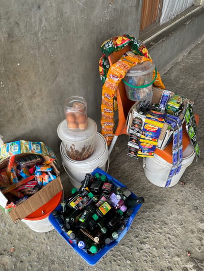 The image depicts an outdoor vendor setup in Ikorodu, Nigeria, featuring various packaged goods and food items displayed on the ground and elevated on buckets and a plastic chair. No individuals are present in the scene.
On the left foreground, a white plastic bucket with a partially visible orange lid supports a cardboard box filled with assorted individually wrapped snacks, including "Krim Kracker," "Chocolup," and "Jam Crackers." Adjacent to this, a blue plastic crate contains numerous small, dark-colored glass bottles with various cap colors (green, black, yellow, purple), some labeled "Action Bitters."
Against the grey concrete wall, two white plastic buckets are stacked. The top bucket supports a clear plastic container holding approximately five brown eggs. Inside the top bucket, beneath the egg container, a viscous brown substance is visible.
To the right, an orange plastic chair serves as a display stand. Long strings of "Classic Peanut Burger" snacks are draped over the chair. On the seat of the chair, a clear plastic bucket with a green handle holds additional packaged items. Also on the chair are numerous sachets and small packets of goods, including "Checkers Custard Vanilla" (yellow), "Seaman's" (blue), "K1 Klicker" (green), and dark-colored sachets, possibly "Gereen Bitters." This entire arrangement rests on another white plastic bucket.
The ground is a rough, dirty concrete surface with minor debris. The background wall is a textured, light grey concrete, scratched and stained, with faint, illegible markings. Above the right side of the display, a section of a building with wooden paneling and a white curtain or cloth is visible. The scene is illuminated by bright, even natural light.