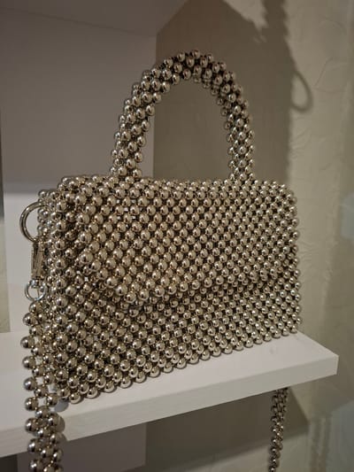 The image shows a close-up of a silver beaded handbag with a top handle and a shoulder strap. The handbag is composed of numerous small, spherical silver beads, creating a textured, metallic surface. The bag is placed on a white shelf, and in the background, a textured wall is visible, with a shadow cast on it. The lighting appears to be from the right, casting a shadow of the handbag and handle onto the wall. The environment suggests an indoor setting, possibly a retail display or a home shelf. The image does not contain any information or context related to the city of Korenovsk, Russia.