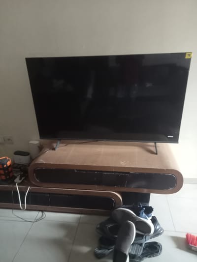 The image displays a living room scene in Gwarinpa, Nigeria. A large, black flat-screen television is mounted on a modern, light brown wooden entertainment center with curved edges and dark drawers. The entertainment center sits on a light-colored tiled floor. To the left of the entertainment center, on the floor, are several electronic devices, including an orange and black portable speaker, a white cylindrical object, and a small white device. A white power cord snakes across the floor from the entertainment center. Underneath the entertainment center, a pile of shoes is visible, including black sneakers and gray slip-on shoes. A red-bristled broom is partially visible on the right side of the frame. The walls are painted a light, neutral color.