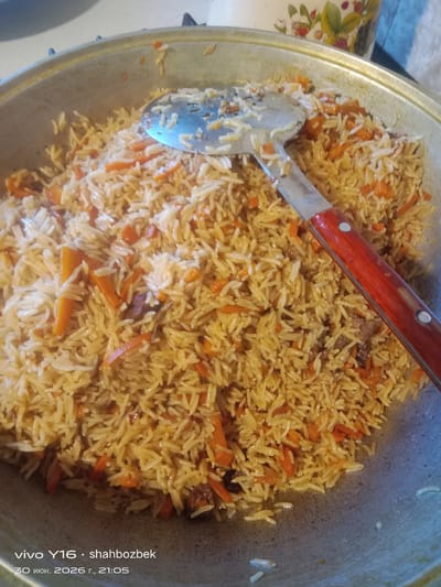 This media file is a close-up, top-down shot of a large pot filled with pilaf, a rice dish. The pilaf appears to be a traditional Central Asian style, featuring long-grain rice mixed with julienned carrots and small pieces of meat. A metal serving utensil with a wooden handle is partially submerged in the pilaf, suggesting it is being served or has just been prepared. The setting is a domestic one, likely a kitchen, indicated by the pot and the background which shows a blurred object with a floral pattern and a glimpse of a stovetop. The lighting is warm and appears to be indoor lighting, suggesting it is not daytime.
Visible text in the lower-left corner of the image indicates it was taken on June 30, 2026, at 21:05, with the phone model "vivo Y16" and the username "shahbozbek". Given the location context of Ufa, Russia, the time of day (21:05) would be evening, and the date (June 30) falls within summer. The overall impression is of a hearty, home-cooked meal.