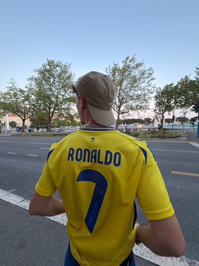A person wearing a yellow Cristiano Ronaldo soccer jersey and a tan baseball cap is standing on a gray asphalt road. The jersey has the name "RONALDO" and the number "7" in blue lettering on the back. The person is positioned with their back facing the camera, and the left side of their face is partially visible under the cap. The background features trees, buildings, and a marina with boats. A traffic light is visible in the distance. The scene is located in Grad Rijeka, Croatia.