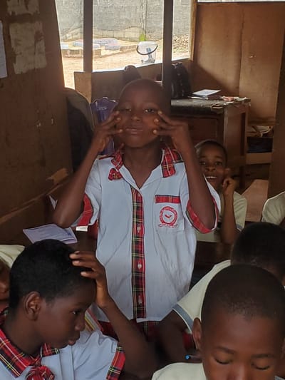 A classroom setting in Ukwu-Abwa, Nigeria, depicts a group of students in school uniforms. The uniforms are predominantly white with red and green plaid accents, including a patch on the chest. One student is actively engaging with the camera, holding their hands to their face. Other students are seated, some with their heads resting on their hands. The room has a rustic appearance with visible exterior elements like windows, showing parts of the surrounding area. The scene appears to be taken in the afternoon or early morning, with natural light illuminating the classroom.
