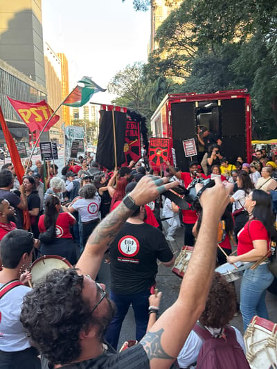 A street-level, medium shot depicts a political demonstration in São Paulo, Brazil. Numerous individuals, primarily dressed in red and black, are gathered on the street. Some are beating drums, while others hold signs and flags. One flag is red with the letters "PST" written on it, while another is green, white, and orange. One man in the foreground is holding a camera and taking photos. The overall mood is energetic and determined. A truck is parked on the left of the frame. Buildings and trees are visible in the background.