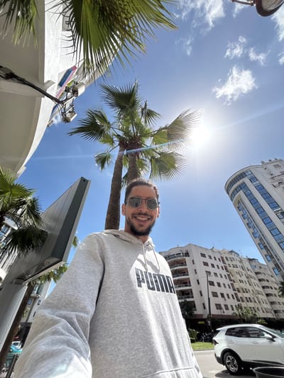 The image depicts a male individual captured in a low-angle selfie format, smiling and wearing reflective sunglasses and a grey "PUMA" brand hoodie. The scene is set outdoors under a bright blue sky with scattered white clouds, featuring a prominent sun glare in the upper right quadrant.
The environment is an urban setting in Tanger, Morocco. Multiple tall palm trees with green fronds are visible, framing the sky and partially obscuring architectural elements. On the left, a modern, white building with mounted technical equipment and cables is partially visible, alongside a distinctive grey, angular architectural structure. On the right, a prominent, multi-story building with a rounded facade and numerous blue-tinted windows rises upwards. Further back, a large, multi-story white residential building with balconies is present. A white SUV is visible on a street or roadside in the lower right background, behind the individual, next to a landscaped area featuring grass and additional trees. The overall impression is a sunny day in a contemporary city district.
