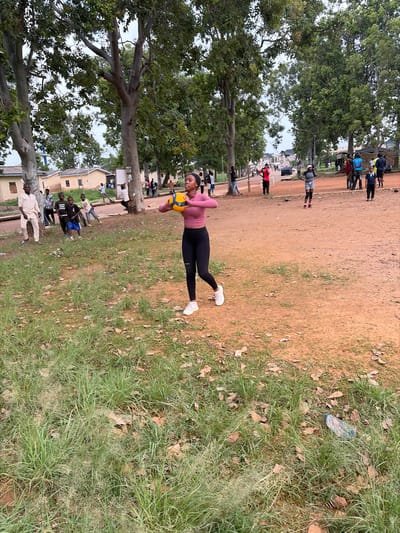 The image depicts an outdoor scene in Rafin Par, Nigeria. A group of people are gathered, suggesting a social or sporting event. The main subject is a young woman in the foreground, wearing a pink long-sleeved top and black leggings, with white sneakers. She is holding a yellow and blue volleyball and appears to be in motion, possibly during a game or practice. Her body is angled, and she is looking down towards the ball, her expression unreadable.
Behind her, a patch of dry, dusty ground gives way to a grassy area with scattered leaves. In the background, more people are visible, some standing in small groups, others walking. There are several buildings with light-colored walls and darker roofs, indicative of a residential or institutional area. Tall trees with dense green foliage are scattered throughout the scene, providing shade. The overall lighting suggests it is daytime, possibly late afternoon given the warm, somewhat diffused light. The presence of volleyball suggests a casual sports activity. The setting appears to be a community space or school grounds. No visible text is present in the image.