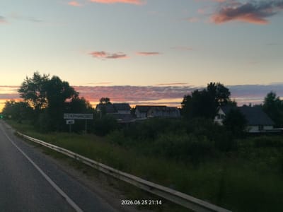 This media file shows a rural scene at dusk. The sky is painted with soft pink and orange clouds against a pale blue backdrop, indicating either sunrise or sunset. In the foreground, a paved road runs along the left side of the frame, bordered by a metal guardrail. Lush green vegetation lines the roadside and extends into the background.

Dotted among the trees and greenery are several houses, suggesting a village or settlement. One prominent signpost on the right side of the road displays text in Cyrillic. The date and time stamp at the bottom right corner reads "2026.06.25 21:14," confirming it is late evening. The overall mood is serene and peaceful, characteristic of a quiet rural evening. The presence of the road and the settlement implies this is likely a view from a vehicle traveling through the area. The location is identified as Starnikovo, Russia.
