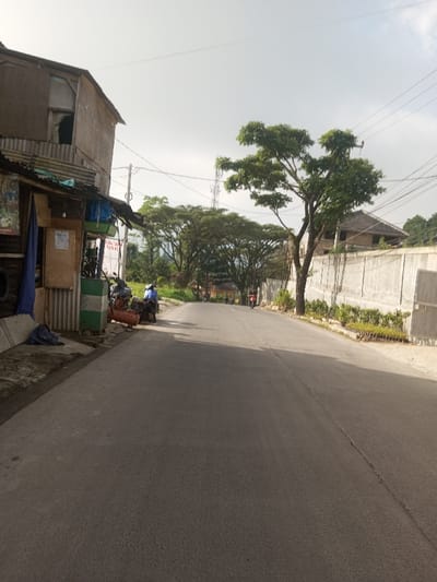 A quiet, paved road winds through a residential area. A small shop with a corrugated metal awning sits on the left, with a motorcycle parked nearby. Across the road is a tall concrete wall lined with green plants. Trees canopy the road further down, creating a tunnel-like effect. Several people on motorbikes are seen in the distance. Power lines stretch across the sky. A tall communications tower is visible in the background. The sky is overcast.