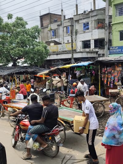 This image depicts a bustling street scene in Benapole, Bangladesh, likely a market area given the presence of numerous stalls and vendors. The foreground features a red motorcycle with two individuals riding on it, one driving and the other as a passenger, with a bag of produce hanging from the front. A man in a white t-shirt with a black stripe down the side is walking towards the left, carrying a cardboard box. To the right, a woman wearing a blue and purple headscarf and a red garment is visible. In the background, several market stalls are set up, displaying various goods, including what appear to be packaged items and food. People are seen browsing and interacting within the market. The environment appears to be an outdoor urban setting with buildings lining the street and overhead wires present. The overall atmosphere suggests a lively commercial activity.