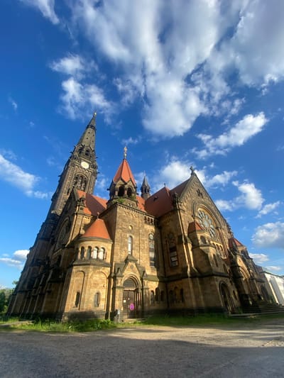 The image depicts the Zionskirche in Dresden, Germany. The architectural style appears to be Gothic Revival. The church is constructed of stone, with red tile roofing. The facade features a tall spire, a large rose window, and other windows of varying sizes. The sky behind the church is a bright blue with scattered white clouds. To the right of the church is a paved area, possibly a road or walkway. There are no people visible in the frame. The image has been rotated.