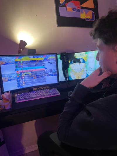 The image displays an indoor scene, likely a private residence, featuring a gaming setup. No specific visual information within the image allows for pinpointing a precise location within Plaisir, France.

A young male individual, with short, dark, curly hair and wearing a dark long-sleeved garment, is seated at a dark-colored desk, viewed from the right side. His right hand is raised, with the thumb supporting his chin and fingers resting on his cheek, indicating a contemplative or focused posture as he observes the dual monitor display.

The desk supports two computer monitors. The left monitor displays a video game interface, specifically a scoreboard for a racing game, listing character names such as "Maskass blanc," "Yoshi," and "Bowser" with numerical scores or positions. A dark webcam is mounted centrally on top of this monitor. The right monitor exhibits visible screen damage, characterized by faint vertical lines, and displays a desktop background featuring the smiling anime character Naruto Uzumaki. Dark over-ear headphones are draped over the top right corner of the right monitor.

A mechanical gaming keyboard with multi-color RGB backlighting (displaying pink and purple hues) is positioned on the desk in front of the monitors. To the left of the keyboard, a white paper cup with orange and brown geometric patterns is present.

The room is dimly lit, with primary illumination originating from the monitor screens and a wall-mounted lamp located in the upper left. This lamp, featuring a reddish-brown conical shade, casts a warm, upward-directed light onto the light-colored wall. A framed piece of abstract art, composed of colorful geometric shapes (red, purple, orange, yellow) on a dark background, hangs on the wall above the right monitor.