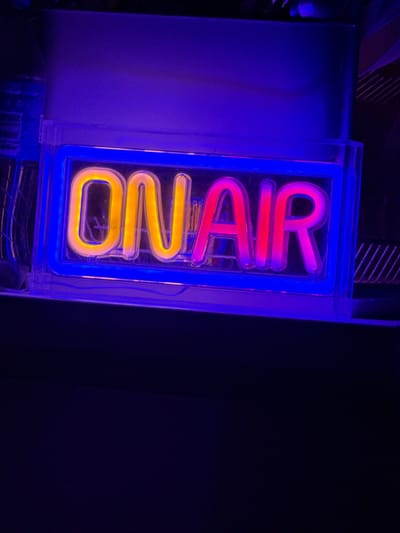An "ON AIR" neon sign is illuminated with yellow and pink lights, encased in a clear plastic box with a blue neon border. The sign is positioned in a dimly lit environment, likely indoors, with a dark background. The upper portion of the image shows a purple-hued surface, possibly a wall or ceiling. The sign itself is the primary subject, with the bright neon lights contrasting against the darker surroundings. The image does not contain any information about the city of Drianovets, Bulgaria, or any people.