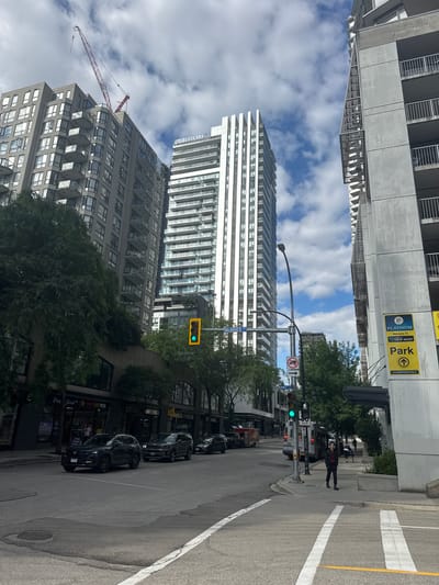 This image captures a street scene in New Westminster, Canada, featuring a combination of modern architecture and urban infrastructure. Dominating the skyline are several high-rise buildings, including a residential complex with balconies and a tall, sleek white building. A construction crane is visible extending from behind one of the buildings, indicating ongoing development.
The street itself is paved and marked with pedestrian crosswalks. Traffic lights are active, showing a green signal. Several vehicles are present, including dark-colored SUVs and sedans on the road, and a bus is stopped at the side. Pedestrians are also visible: one individual in a red and black jacket is walking on the sidewalk, and another person is seen further down the street with a dog.
Signage is present, including a "Park" sign with the "Platinum Parking" logo and an arrow indicating direction, as well as street signs identifying "McINNES ST". The environment is characterized by trees lining the sidewalks and a partly cloudy sky. The overall impression is that of a bustling urban environment in daylight.