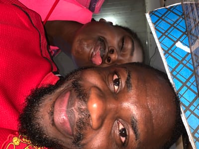 This is a close-up, overhead shot of two Black males in what appears to be a bedroom in New Stadium, Nigeria. The lighting is dim, suggesting an indoor setting. The male in the foreground, to the left of the image, is positioned looking up, with his mouth slightly open and his tongue extended. He has a beard and dark, curly hair. He is wearing a red shirt with black trim. The male behind him, to the right, is also looking up and appears to be asleep or unconscious, with his eyes closed and his mouth slightly agape. He is wearing a bright pink garment. A blue and black checkered notebook is visible to the far right of the image, partially obscuring the background. The overall impression is one of intimacy or possibly distress, given the unusual positioning and expressions of the individuals.