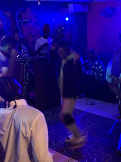 The image depicts an indoor scene, likely a club or party venue, illuminated by vibrant blue lighting. In the foreground, a person wearing a white shirt is partially visible from the back, with their head turned away from the camera. In the midground, several individuals are present. One person, wearing a red beanie and a white t-shirt, appears to be interacting with a DJ setup. Another individual, wearing a black and white checkered shirt, is seated or leaning. The background features a mural on the wall, stylized with faces and sunglasses. The floor is tiled and reflects the blue light. The overall atmosphere suggests a lively social gathering. No specific details about the city of Durumi, Nigeria, are discernible from the image itself.