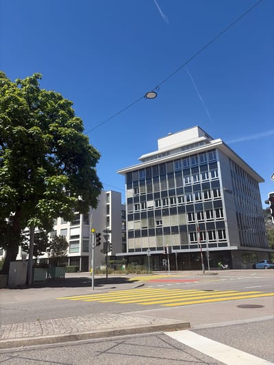 In Zurich, Switzerland, on a clear, sunny day, a street scene is captured. The foreground features a pedestrian crossing with bright yellow stripes on the asphalt. To the left of the crossing, a large, leafy green tree stands tall, its branches extending towards a clear blue sky. Behind the tree, a modern white building with multiple windows is visible. To the right of the crossing, a multi-story office building with a dark facade and a grid of windows dominates the view. The building's architecture is distinctly mid-century modern, with a white roofline. A traffic light, displaying red and yellow signals, stands at the intersection. The street is paved with asphalt, and there are sidewalks made of cobblestones and concrete. A few vehicles are present, including a blue car parked on the right side of the street and a green bicycle leaning against a wall near the office building entrance. A sign with the letter "R" is visible on the glass of the office building. The overall environment suggests an urban setting with a mix of residential and commercial buildings, characterized by clean lines and orderly infrastructure.