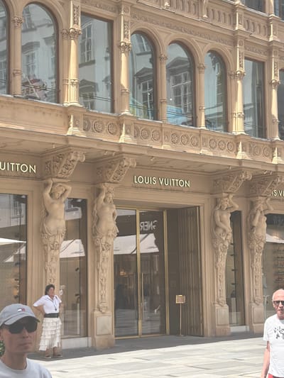 This media file captures a close-up, slightly angled view of the Louis Vuitton store facade in Vienna, Austria. The building's architectural style is ornate, featuring classical details like arches, decorative carvings, and sculptures of reclining figures adorning the upper levels. The facade is primarily light beige stone.
The activity taking place is not explicitly clear, but the presence of people suggests it's an active storefront, likely during business hours. One person, a man wearing a white t-shirt and sunglasses, is partially visible in the upper right corner, looking towards the camera. Another person, wearing a white ruffled skirt and white top, is standing near the entrance on the right side of the frame, looking away. A third person's head and shoulders are visible in the bottom right, wearing a grey baseball cap and sunglasses, also looking away.
Notable details include the large arched windows on the lower floors, which reflect the surroundings, and the prominent "LOUIS VUITTON" text in gold lettering on the facade. The lighting suggests it's daytime, possibly a sunny day given the reflections. The overall impression is one of a luxury retail environment combined with historical architecture.