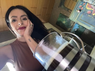 A woman with long dark hair and wearing a brown top poses for a selfie with a glass mug, possibly filled with coffee, held at an angle above her. She rests her face on her hand, which features a ring on one of her fingers and a tattoo on her arm. The mug is striped with black and silver bands. In the background, a painted cabinet is visible along with some landscape paintings. The location in the city of Konz, Germany is not discernible from the provided image.