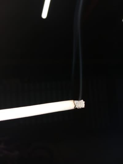 A close-up, horizontal shot focuses on a lit cigarette against a black background. The cigarette, held horizontally, is positioned in the lower left quadrant of the frame. Its white paper body is visible, with the burning tip showing ash and a faint orange glow. Thin wisps of white smoke curl upwards from the tip, ascending into the darkness. The background is uniformly black, suggesting a dimly lit or nighttime environment. There is no indication of the city of Dinskaya, Russia, or any urban setting within the image.