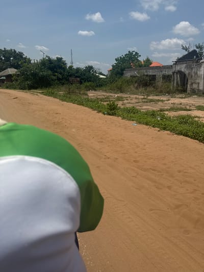 The image presents a view of a sandy area or path in Ihiagwa, Nigeria, with vegetation and trees lining one side. An aged, weathered building is visible in the background. A partially visible object in the foreground appears to be green and white. The sky is blue with scattered clouds. There is a tall communications tower visible in the background to the left.