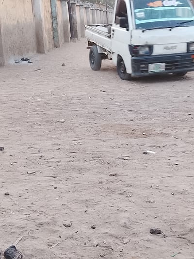 The image captures an outdoor scene, likely on an unpaved road or dusty open area, during what appears to be daytime. The ground is predominantly a light brown or tan color, consisting of dirt or sand, scattered with numerous small rocks, debris, and what resembles dried plant matter or litter.
A small, white pickup truck is prominently featured on the right side of the frame, positioned as if it is either moving slowly, pulling away, or has just stopped. The truck has a flatbed cargo area at the back. Its front displays headlights, a dark-colored grille, and a bumper. The brand name "HIJET" is visible on the front grille area. A license plate, with green alphanumeric characters on a white background, is affixed to the front bumper, although the characters are not fully legible due to blur and resolution. There is also a sticker or decal on the upper part of the windshield, with an orange element and some text that is too blurry to read distinctly. The truck's wheels are visible, with the front wheel showing a slight blur, suggesting motion.
In the background, on the left side, there is a low wall or fence-like structure composed of light-colored, possibly mud or concrete, blocks or pillars, indicating the boundary of a property or compound. The general setting suggests a rural or semi-urban environment. No people are visible in the image, and no specific activity beyond the presence of the truck is apparent. The lighting is soft and even, characteristic of daylight hours, without harsh shadows.
