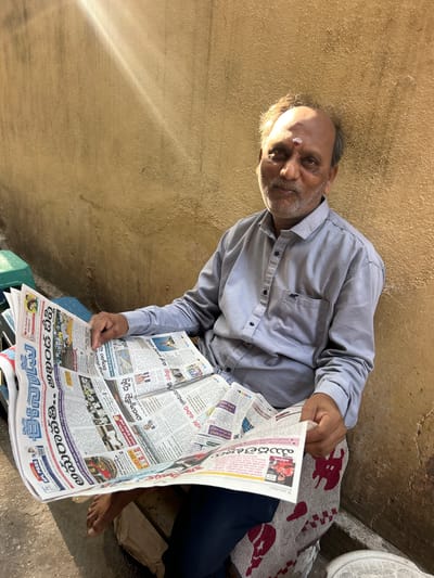 An elderly South Asian man in Puttaparthi, India, sits outside reading a Telugu newspaper. He wears a light blue long-sleeved shirt, dark blue pants, and a white tilak on his forehead. The newspaper is spread out in front of him, resting on his lap and a green container. He sits on the ground next to a blanket with red patterns. The background consists of a rough, tan-colored wall. The light indicates it is likely daytime.