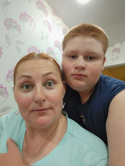 This is a selfie taken indoors, possibly in a home in Markovskii, Russia. Two individuals are visible: a woman in the foreground and a young person behind her. The woman has auburn hair, is wearing a light blue t-shirt, and has earrings and a necklace. The young person has short, reddish-blond hair and is wearing a dark blue t-shirt with a cross pendant. They are positioned close together, with the woman's head and shoulders filling the left side of the frame and the young person's head and upper body filling the right. The background is wallpapered with a light-colored pattern of pink and grey flowers. The lighting appears to be artificial, coming from above.