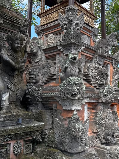 This image captures a detailed view of ornate stone carvings and architecture at a temple in Ubud District, Indonesia. The foreground features a large, intricately carved stone statue of a warrior or deity, adorned with a headdress and detailed clothing. To its right, another stone figure with a similar headdress is partially visible. Behind these figures, the temple structure is evident with multiple levels of carved stonework and red brick walls. Several grotesque or demonic-looking faces with bared teeth are incorporated into the architecture, some appearing as gargoyles or decorative elements. A winged creature, possibly a mythical bird or demon, is perched on a higher level, also intricately carved. The background shows glimpses of trees and a blue sky, indicating an outdoor setting. The overall scene conveys a sense of ancient artistry and religious significance.