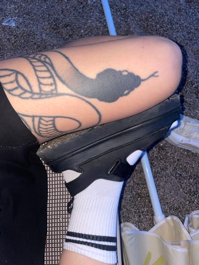 The image presents an overhead view focusing on a person's leg with a prominent snake tattoo. The tattoo depicts a stylized serpent with a distinct head and scaled body. A black sandal is positioned partially on the leg, obscuring part of the tattoo. The person wears a white sock with three black stripes at the top. Other details include a glimpse of a white shoe, an umbrella support pole, and a woven mat surface.
The image was not taken in Rafailovići, Montenegro.