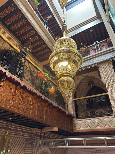 The image presents an upward view within an ornately decorated interior space, characteristic of traditional Moroccan architecture, likely a riad or courtyard in Marrakesh.
A large, multi-tiered golden chandelier with intricate filigree designs dominates the center, glowing warmly and illuminating its surroundings.
The architecture features various traditional elements. Walls are constructed from bricks and textured plaster, with sections adorned by geometric Zellige-style tiling in shades of blue, green, and beige. Multiple levels of balconies are visible, featuring dark wooden panels with painted or carved floral and geometric motifs, protected by decorative wrought iron railings. Small green plants in terracotta pots are placed on some balconies. A prominent carved wooden balustrade or beam in the foreground displays highly detailed, swirling patterns. Brick arches frame windows or doorways on upper levels.
No people are visible, and no specific activity is taking place; the focus is entirely on the architectural details and decorative elements. The scene is warmly lit, primarily by the glowing chandelier, complemented by ambient light from above.
On the far right, a silver-colored metal structure stands next to a banner with Arabic text. The text reads: "تحت شعار" (Under the slogan) followed by "المسيرة الخضراء" (The Green March), referencing a significant historical event in Morocco.
The overall impression is one of rich cultural heritage, intricate craftsmanship, and opulent design.