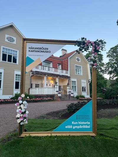 In Vantaa, Finland, at Hakansbolen Manor Museum (Hakansbolen Kartanomuseo), a large, ornate golden frame is placed on a grassy lawn in front of the museum building. The frame is decorated with artificial pink and white roses and greenery. The museum is a light beige, two-story building with white trim, a red tiled roof, and several windows. A porch with white railings and steps leads to the entrance. The frame contains a sign with the museum's name and website, "hakansbolen-kartano.fi," along with social media icons and the Finnish text "Kun historia elää ympärilläsi," which translates to "When history lives around you." The sky is clear and blue, and trees are visible in the background. The ground around the frame and in front of the museum is covered with gravel and grass, with flower beds lining the path. There are no people visible in the image.