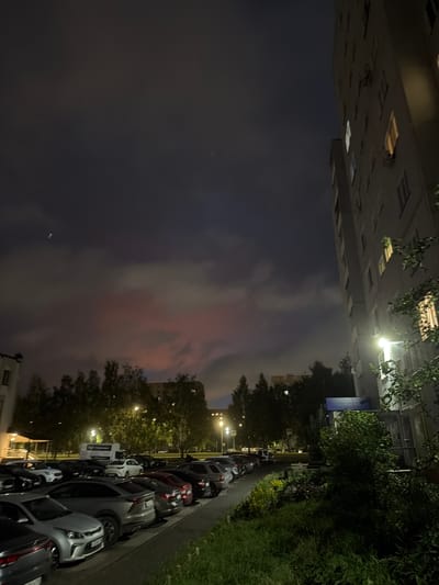 This image captures a street scene at dusk in Nizhnekamsk, Russia. The perspective is rotated 90 degrees clockwise. On the left, a tall, multi-story building dominates the frame, its windows illuminated from within, suggesting occupancy. Directly opposite, a row of parked cars lines the street. The cars are of various models and colors, primarily dark grays, silver, and white. Some of the cars have their headlights on.
Beyond the cars, a sidewalk and a strip of greenery with trees and bushes are visible. Streetlights cast pools of yellow light onto the road and sidewalk, illuminating the scene. The sky above is a mix of dark grays and lighter hues with hints of pink and orange, indicative of the fading daylight. The clouds are wispy, suggesting it's a calm evening.
A signpost with yellow diamond-shaped warning signs is present, and further down the street, another sign with some text is visible but unreadable due to the resolution. The overall mood is quiet and residential, typical of a neighborhood at the end of the day. The lighting suggests it is twilight, likely between 6 PM and 8 PM, depending on the season. The scene appears to be a typical urban residential area.