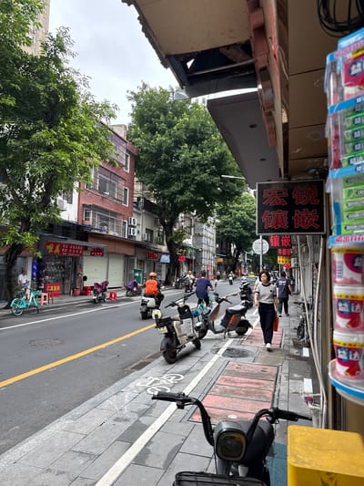 The image depicts a street scene in Liwan District, China. On the right side of the frame, a sidewalk features stacked containers of products, possibly toiletries or cleaning supplies, with Chinese labeling. A woman in a striped shirt and dark pants walks along the sidewalk, carrying an orange bag. Adjacent to the sidewalk is a street with several electric scooters parked and in motion. One scooter, ridden by a person wearing an orange vest and helmet, is moving away from the camera. Another scooter is parked near a store entrance.

The buildings lining the street appear to be a mix of commercial establishments and residential units. Some storefronts have rolled-down metal shutters, while others display signage, including a red sign with Chinese characters. Green trees with lush foliage are present on the left side of the street, with one tree prominently in the foreground. The sky is overcast. The street itself is paved and marked with a yellow line. The overall impression is a busy urban environment.