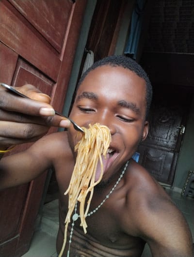 A young Black male is shown from the chest up, looking towards a fork full of spaghetti that he is bringing to his mouth. He is shirtless and wearing a light-colored beaded necklace. The background appears to be indoors, with a dark wooden door visible behind him to the right and a wooden paneling structure on the left. The flooring is a light-colored tile. The scene is consistent with a domestic setting within Ihiagwa, Nigeria.